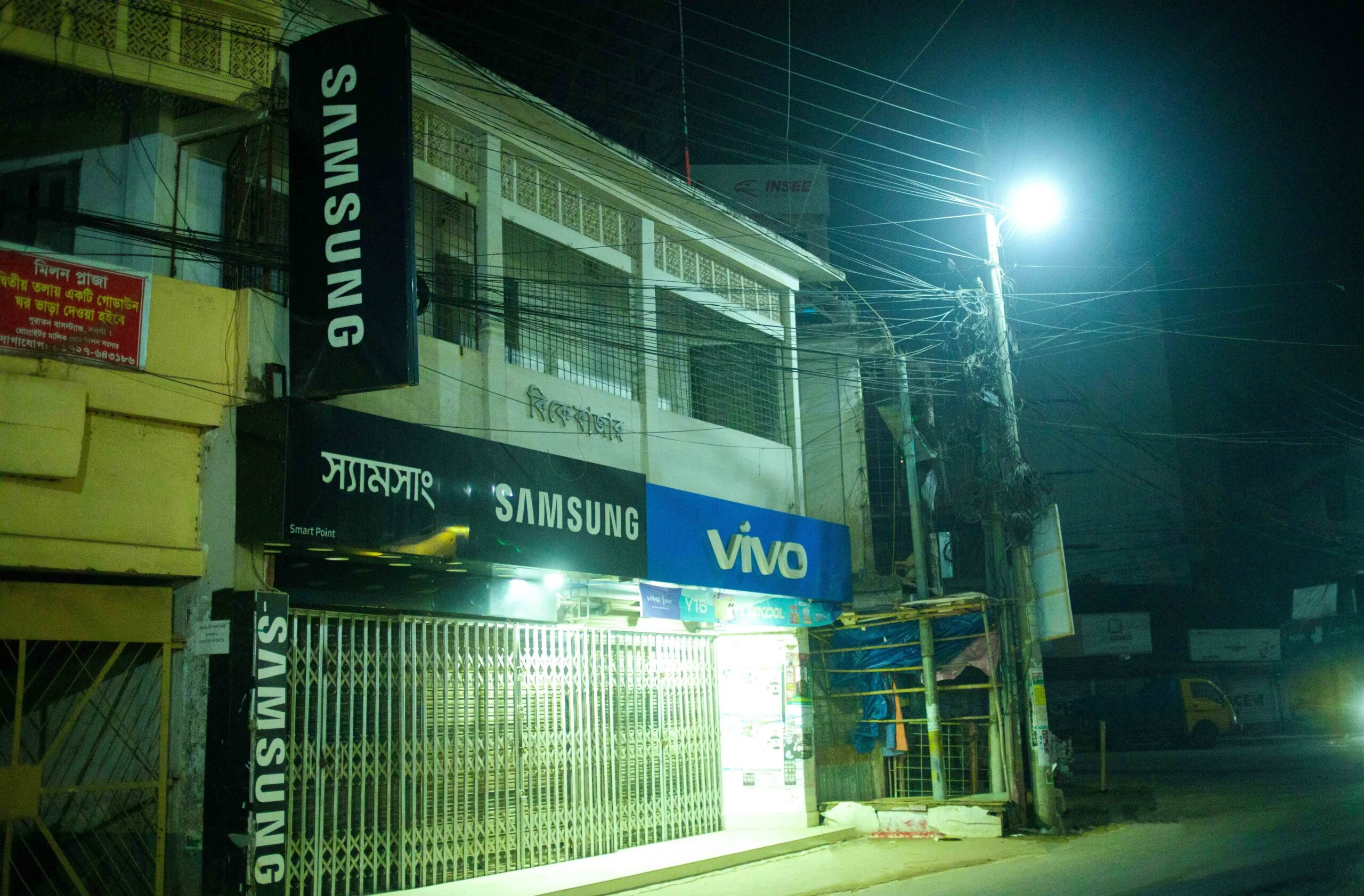 Nighttime view of SAMSUNG and VIVO smartphone stores on Naogaon Main Road.
