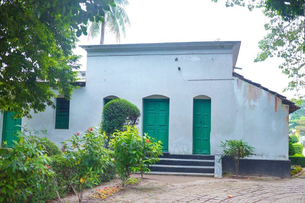 West side view of Patisar Kachari Bari surrounded by trees, featuring two old wooden doors.